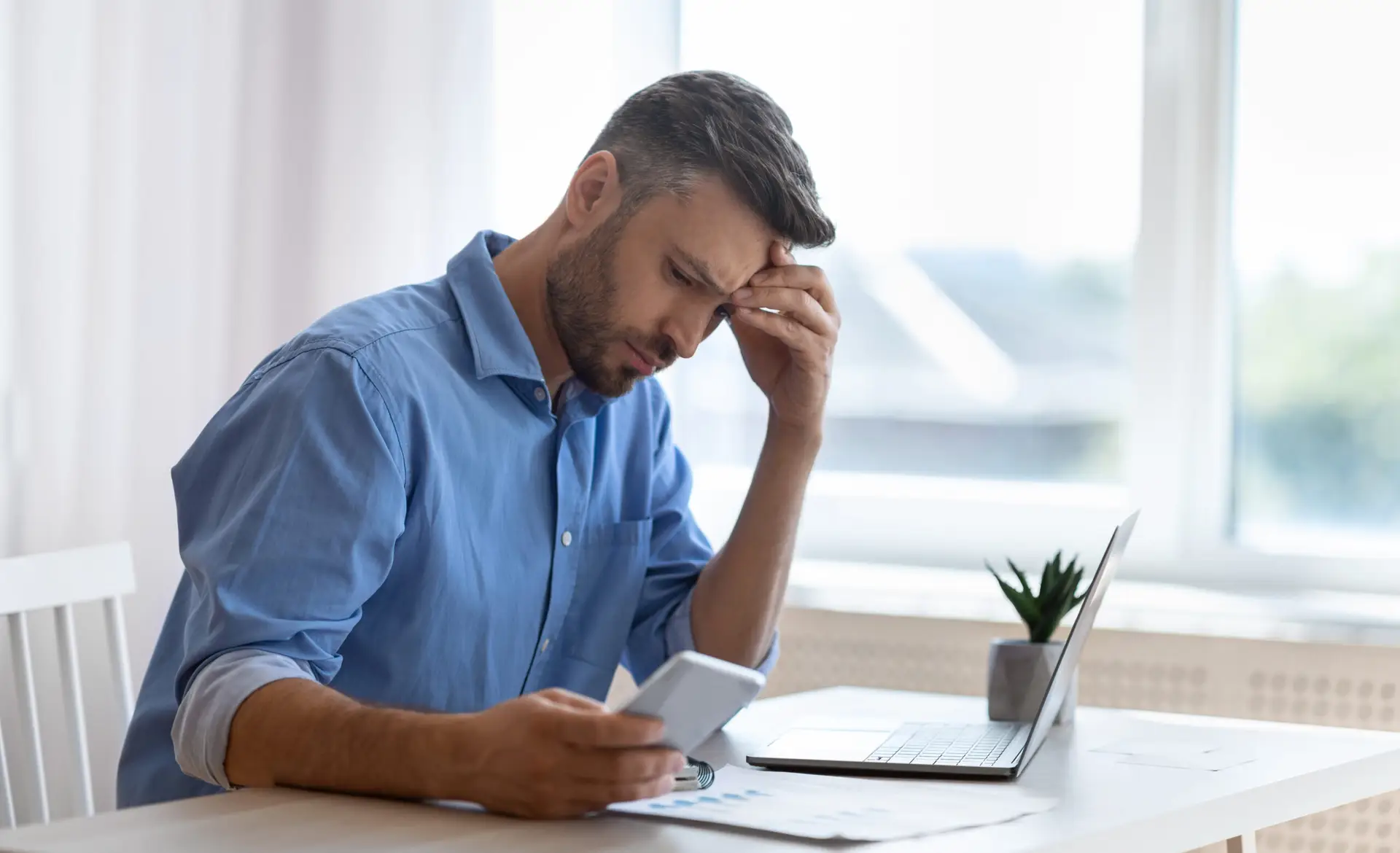 Worried male freelancer looking at smartphone screen while working at home office