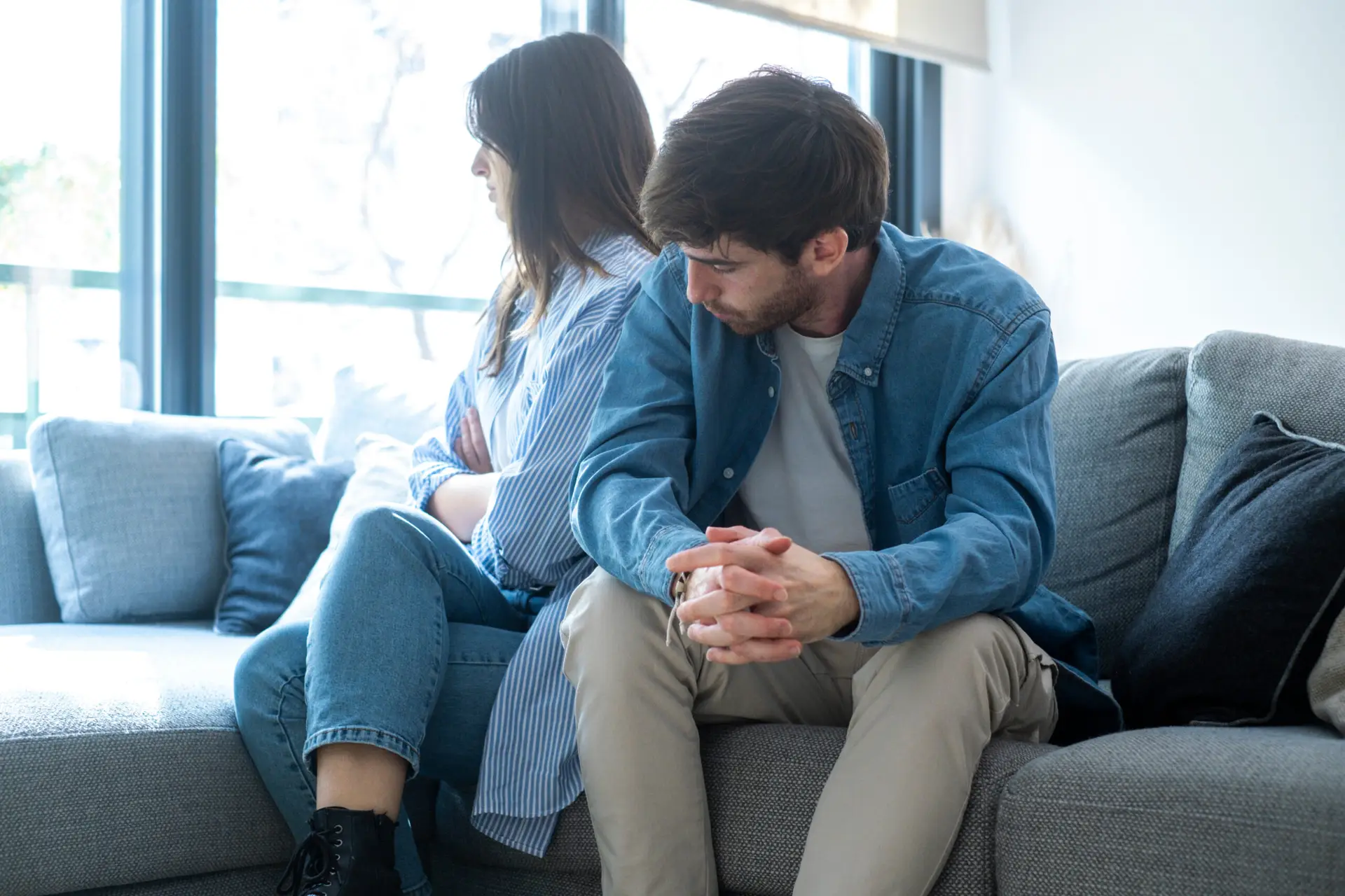 Millennial couple sitting on the couch at home, facing away from each other, struggling with love fading, infidelity suspicions, communication problems, and emotional frustration in their relationship