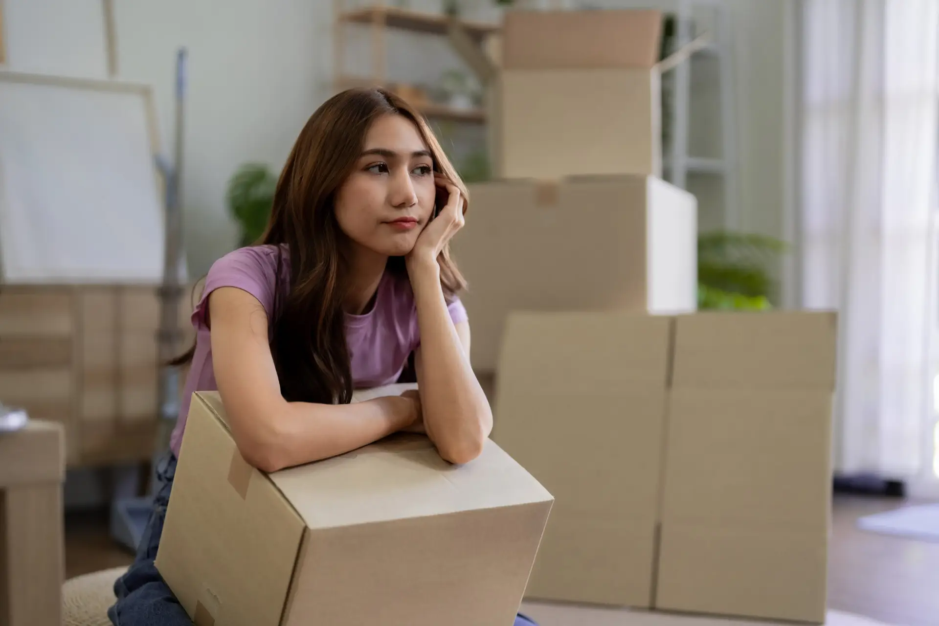 Asian Women Relocating New Home Pondering Next Step During Move In Process  After Moving Day In Living Room With Moving Boxes Near Window