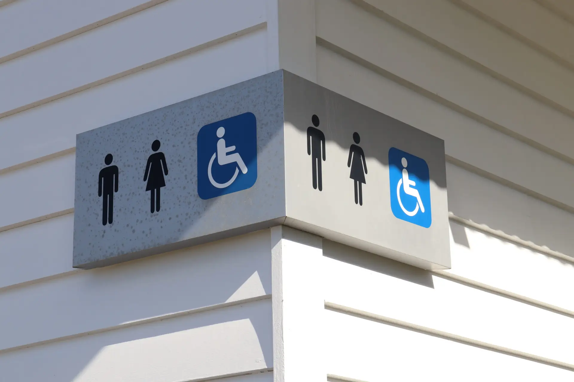 International public restroom sign showing male, female, and accessible icons on a sunlit building corner wall