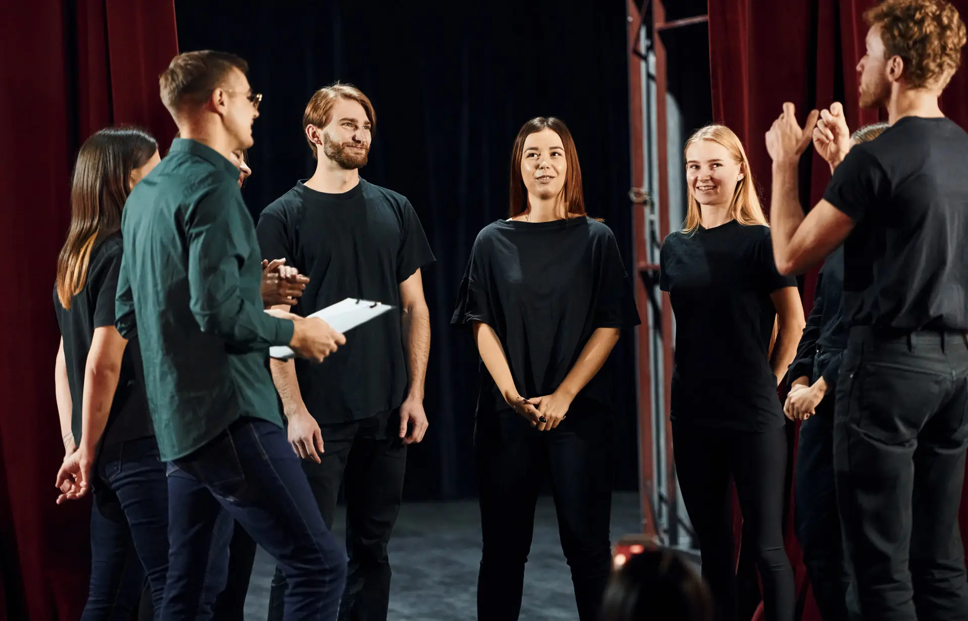 Standing against red curtains. Group of actors in dark colored clothes on rehearsal in the theater