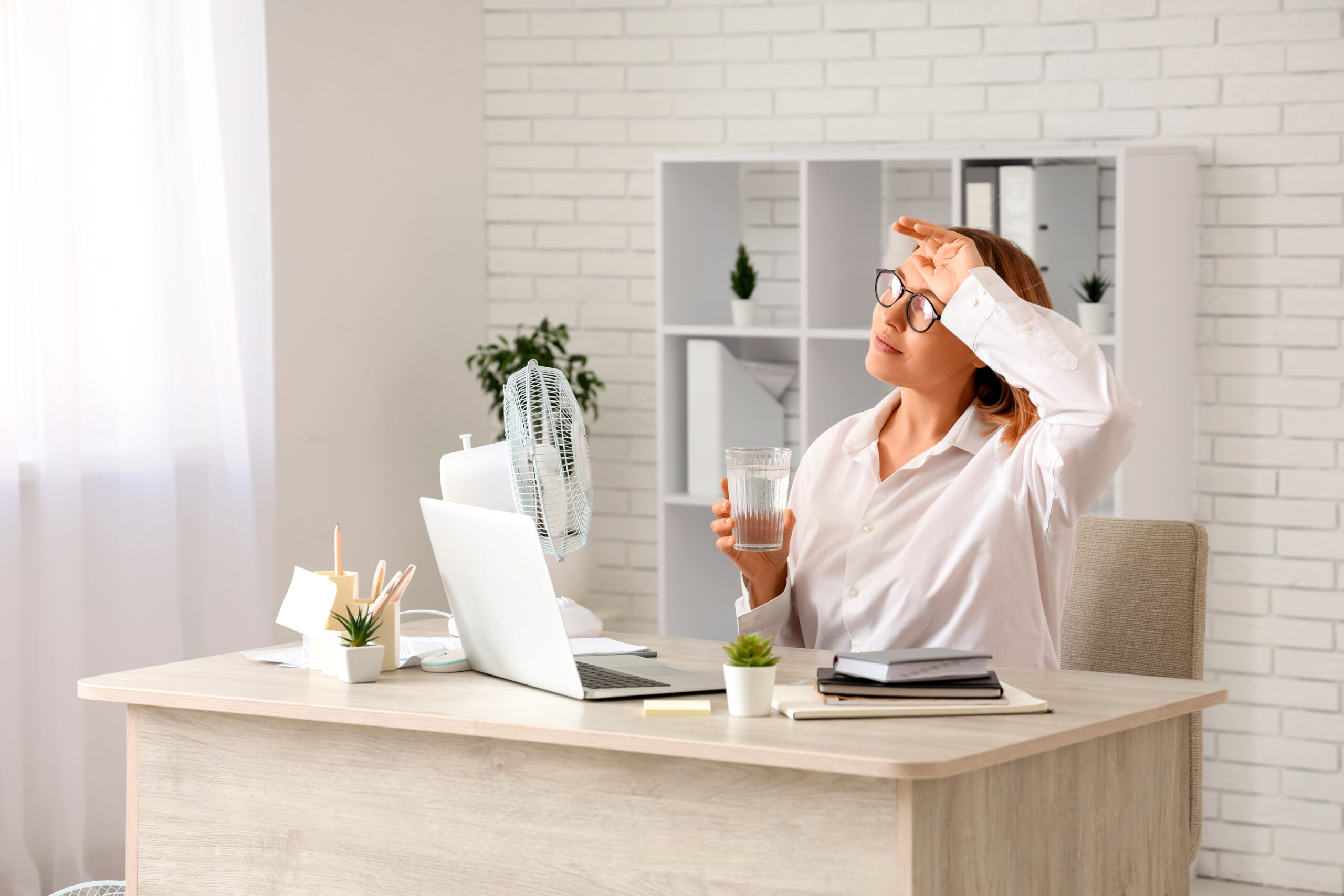 Businesswoman cooling off with electric fan and glass of water while working at desk in hot home office during summer heatwave