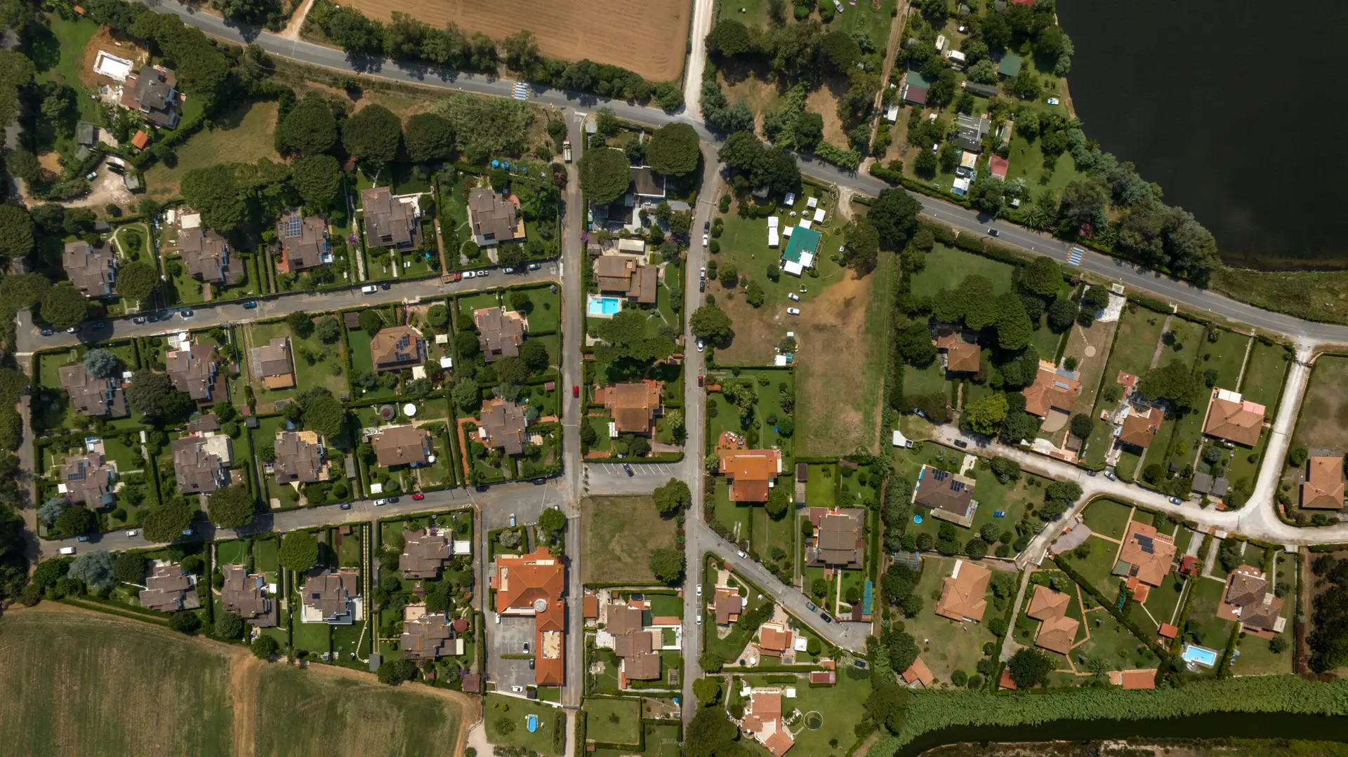Aerial top down view of a quiet residential neighborhood with detached houses, green gardens, private swimming pools and tree lined streets bordering a dark lake, showing suburban layout from above.