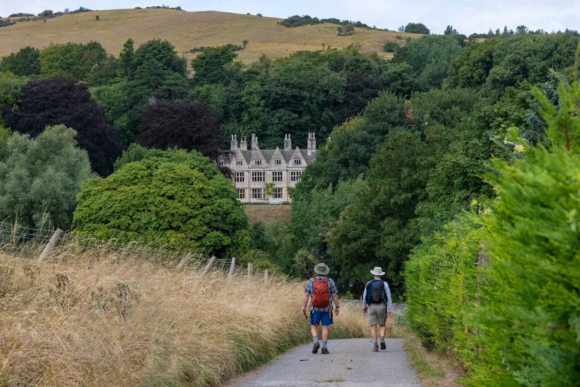 High angle view over downward sloping public footpath Cotswolds Way near Winchcombe, UK with two hikers heading downhill between grasses and trees with traditional buildings between green in distance